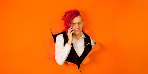Young man with red dreadlocks speaking on smartphone and sticking out of hole of orange background. Stylish hipster in eyeglasses talks on mobile phone.