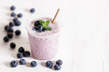 Glass of blueberry milkshake with fresh blueberries on white wooden background.