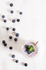 Glass of blueberry milkshake with fresh blueberries on white wooden background.