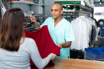 Positive man laundry worker returning clean clothes to customer at dry-cleaning facility