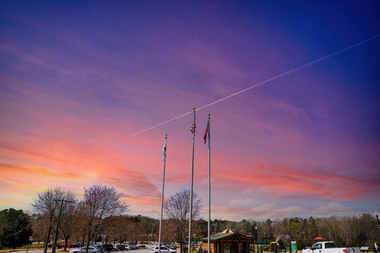 a stunning share of an American flag and two other flags flying on tall flag poles with parked cars, lush green trees with purple sky at sunset at Swift Cantrell Park in Kennesaw Georgia USA
