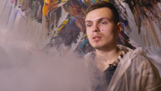 Artist designer draws an eagle on the wall. Craftsman decorator paints a picture with acrylic oil color. Vaper smokes vape. Painter painter dressed in a paint coat. Indoor. Dark magic cinematic look.