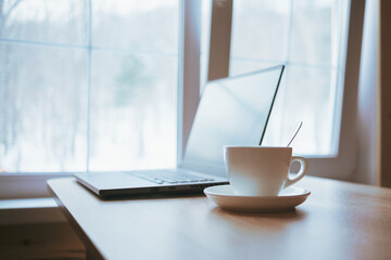 Cup and laptop on the table in front of the window.