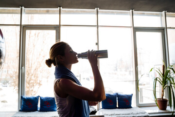 A sporty girl drinks bottled water in the gym in front of a window. Silhouette of a fitness trainer. Sports lifestyle, recuperation in training.