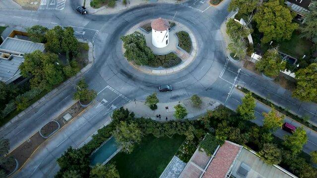 Aerial rising over Leonidas Montes windmill tower in roundabout with cars commuting surrounded by trees, Lo Barnechea, Santiago, Chile