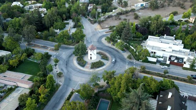 Aerial orbit of Leonidas Montes windmill tower in roundabout with vehicles commuting surrounded by trees, Lo Barnechea, Santiago, Chile