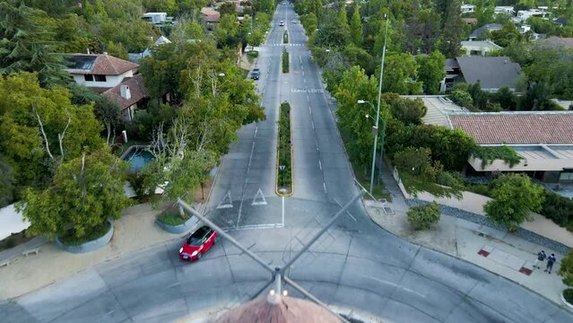 Aerial dolly in of Leonidas Montes windmill in roundabout with cars driving in avenue surrounded by trees, Lo Barnechea, Santiago, Chile