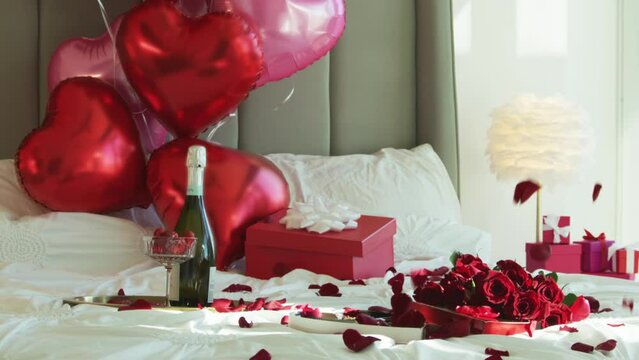 Super slow motion of falling red rose petals on white bed in elegant luxury hotel room with champaign red heart shaped balloons on background. Cinematic romantic background shot on RED camera 6K
