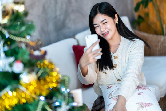 Portrait Cute beautiful positive smile young asian woman using smartphone video call or Selfie at home in the living room indoors New Year holidays.