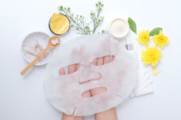 woman hand holding white sheet mask with clay mask wooden spoon yellow flowers and serum skincare...