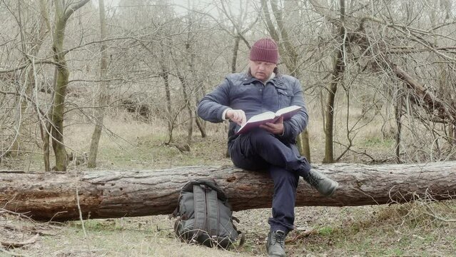 a tourist reads a religious book in the forest sitting on a fallen pine tree