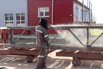 A worker in special clothing is sandblasting a metal building structure at an industrial site. Industrial metal processing.