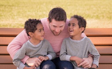 White father is listening to his laughing cute African American twin boys while sitting on bench in Midwestern park - Powered by Adobe