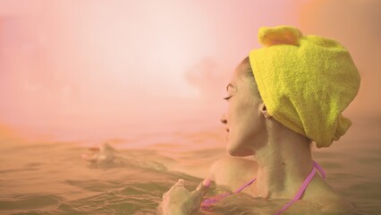Close up caucasian woman in a swim in hot saline mineral water bath at a traditional spa outdoor with soft magic pink lighting and steam. Girl relaxing spring in hot geothermal spa pool is towel cap.