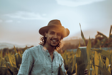 A young hipster farmer inspects the abundance of corn leaves while wearing a hat. An attractive man...