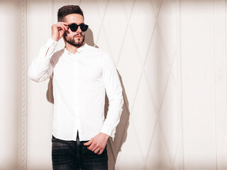 Portrait of handsome confident  model. Sexy stylish man dressed in white shirt and jeans. Fashion hipster male posing near wall in studio lux interior in sunglasses. Window shadow from sun