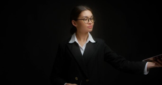 Business lady in glasses and business clothes signs a document attached to a tablet. A strict brunette on an isolated black background signs documents.