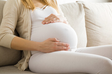 Young adult pregnant woman in white clothes sitting on beige sofa at living room, touching big belly and feeling her baby move. Emotional loving pregnancy time. Baby expectation. Home relax. Side view