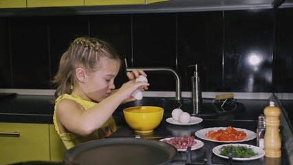 Smart girl learning to cook. Happy and serious young mistress children to cook a Neapolitan egg fried omelette from sausage. Modern Built In Kitchen Appliances in green black color.