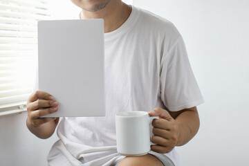 Man in white shirt holding white book and white glass. Mock up of mug and book