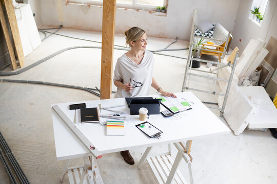 pretty young architect working on construction site in loft and standing by high table wearing glasses and beige top and working on notebook