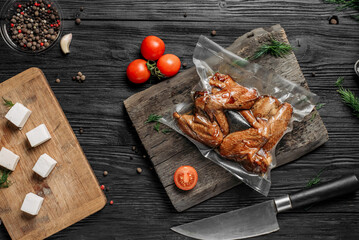 homemade hot-smoked chicken wings, in vacuum packaging on an old wooden board next to vegetables...