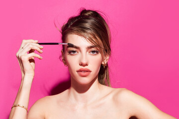 Naklejka premium Combs eyebrows with a brush in a beauty salon. Woman with long eyelashes and thick eyebrows. Woman portrait isolated on pink studio background. Natural beauty brows.