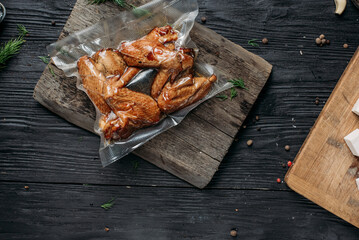 homemade hot-smoked chicken wings, in vacuum packaging on an old wooden board next to vegetables...