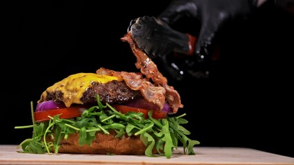 Craft burger is cooking on black background. Consist: sauce, arugula, tomato, red onion, bacon, red currant sauce, ricotta cottage fresh cheese, air bun and marble meat beef. Not made ideal. Looks