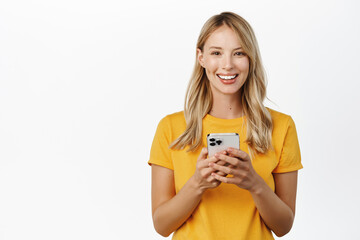 Portrait of smiling young woman, 25 years old, holding smartphone and looking happy, using mobile phone app, standing in yellow t-shirt over white background - 491989343