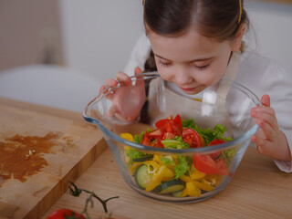 little girl is preparing salad. She is sitting at kitchen table. In front of her variety of...