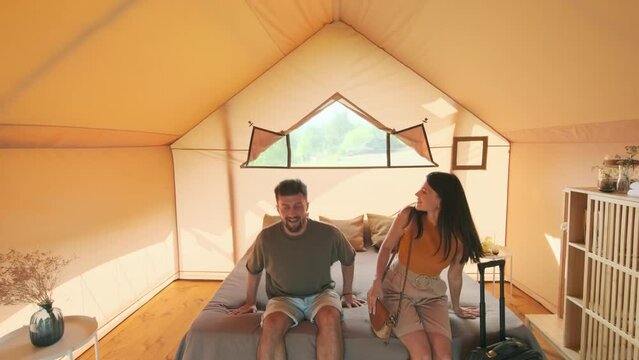 Tracking of young Biracial woman and man carrying luggage, arriving at their tent in glamping, falling down on bed and laughing on sunny summer day