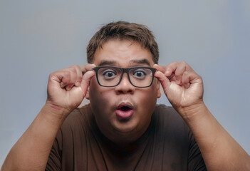 Asian man showing surprise, shock, excitement and hand-holding glasses on a gray background.