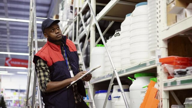 Male supervisor examining goods on shelves with tablet using ladder