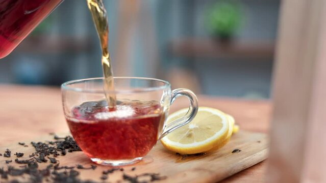 Closeup pouring fresh hot aroma black tea from jar teapot into glass cup on wooden table slowmo