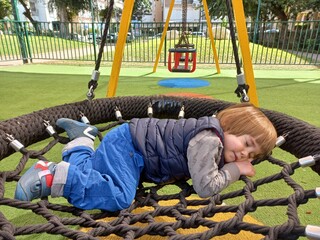 Little cute boy with long blond hair sleeps on a swing in the playground. The child is resting on a...