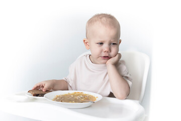 A little boy eats soup with bread on his own in a highchair against the background of a white wall.