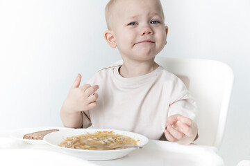 A little boy eats soup with bread on his own in a highchair against the background of a white wall.