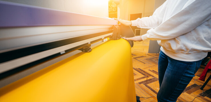 Female hands holding yellow printable material on alarge format printing plotter. Graphic design and  advertising concept.
