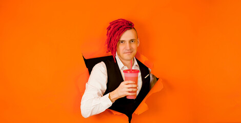 Barman with drink looking through hole in paper background. Male bartender with red dreadlocks and cup of sweet beverage peeking through ripped orange paper background in studio and looking at camera