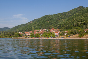 Fototapeta na wymiar Picturesque view on village on shore Danube river. Green forest and hills. Summer day. Travel and holiday. Serbia.