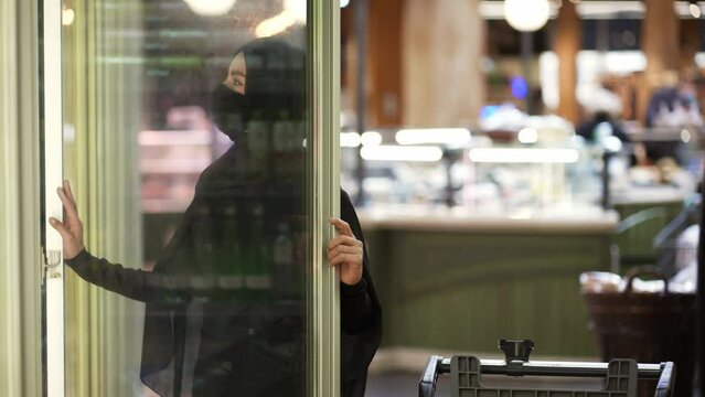 Woman in hijab and protective mask doing shopping, takes product from the freezer shelf