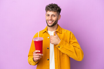 Fototapeta premium Young blonde man holding soda isolated on purple background giving a thumbs up gesture