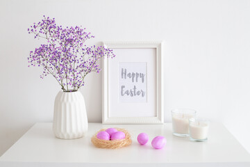 Lilac colored eggs, frame with text HAPPY EASTER, vase with purple gypsophyla flowers, candles and two rabbits made of concrete on white table. Easter home interior decor.