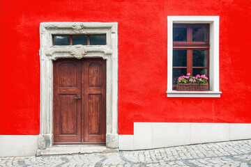 Wooden door home entrance. Window with flower box. Italian architecture background. Vibrant color red wall facade. Small town house exterior. Street of European city building.