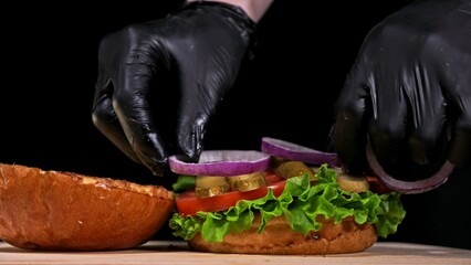 Craft burger is cooking on black background in black food gloves. Consist: sauce, lettuce, tomato, red onion, pickle, cheese, bacon, air bun and marbled meat beef. Not made ideal. Looks real, loving