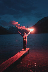 The hiker on the pier lit the emergency red torch and calling for help. Rescue flare and sos signal concept - obrazy, fototapety, plakaty