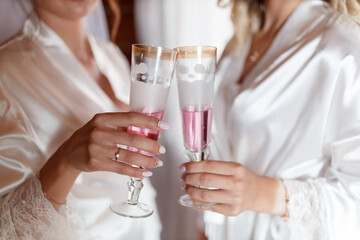 two young girls in white silk robes clink glasses of champagne, close-up without faces. bride's morning in her room