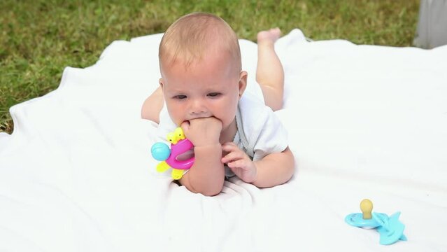 Little newborn girl lying on tummy chewing rattle on blanket, summer day outdoor. Infant having fun outdoors. Infant baby child resting playing learning to crawl. Motherhood teething, babyhood concept