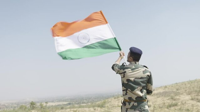 Back view shot of Indian soldier holding waving Indian flag on top of hil - concept of independence or repubilc day celebration, nationalism, victory and freedom
